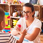 A child and an adult are inside a home environment playing with toy blocks across a table.