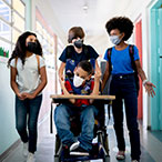 A group of teenagers walk down a school hallway, and one student is being pushed down the hall in his wheelchair. Everyone is wearing facemasks.