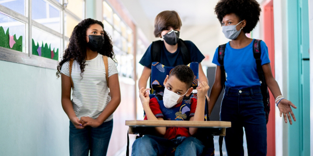 A group of teenagers walk down a school hallway, and one student is being pushed down the hall in his wheelchair. Everyone is wearing facemasks.