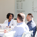 A Black woman wearing a lab coat sits at a table in a conference room speaking to two male colleagues. A whiteboard is visible in the background.