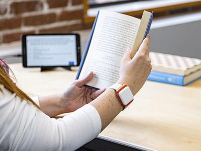 A person wears a Biosensics wrist sensor while reading a hardcover book. The sensor is a white rectangle positioned like a wristwatch.