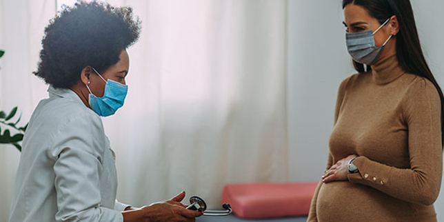 A pregnant woman holds her belly as she sits across from her healthcare provider, who is holding a stethoscope. Both are wearing masks.