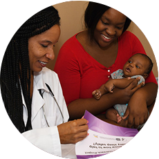 A health care provider wearing a lab coat and a stethoscope reviews a document with a woman holding a baby.